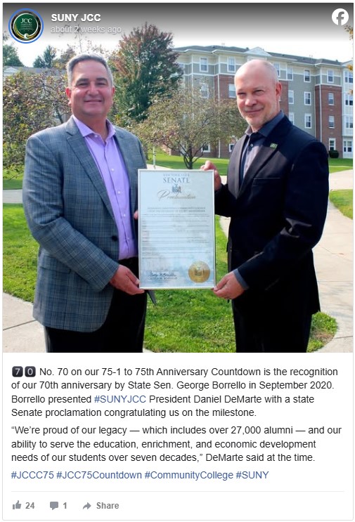 Two men stand outdoors holding a state Senate proclamation. Both are smiling, dressed formally, with a leafy campus and building in the background.